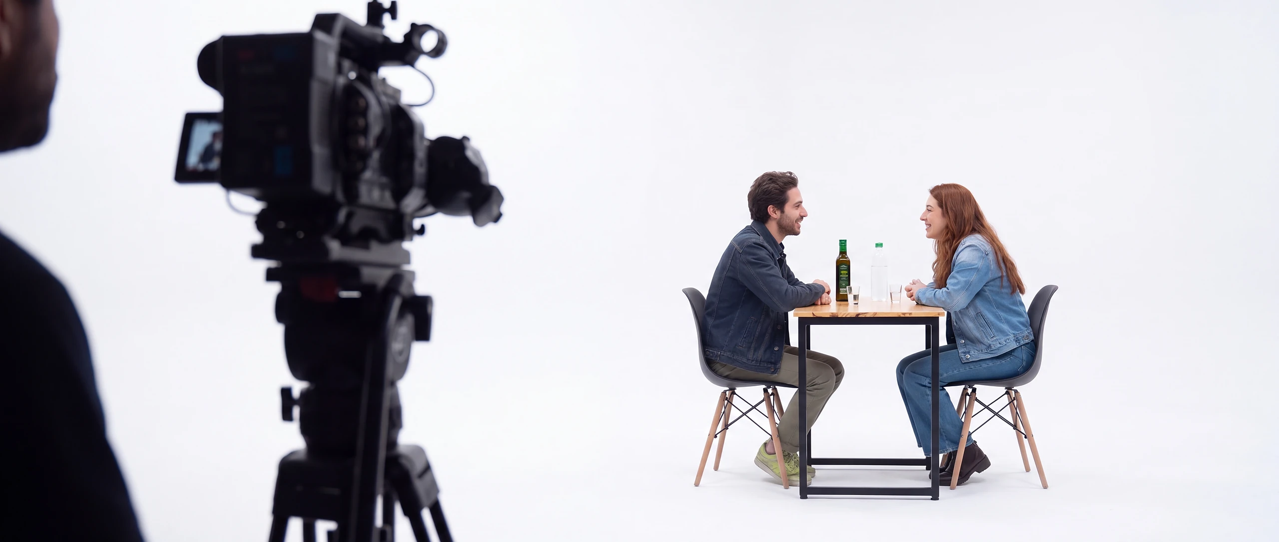 Bastidor ilustrativo do formato: vista por trás de uma câmera de cinema, duas pessoas conversam de frente uma para a outra na mesa de pinho, no estúdio branco.