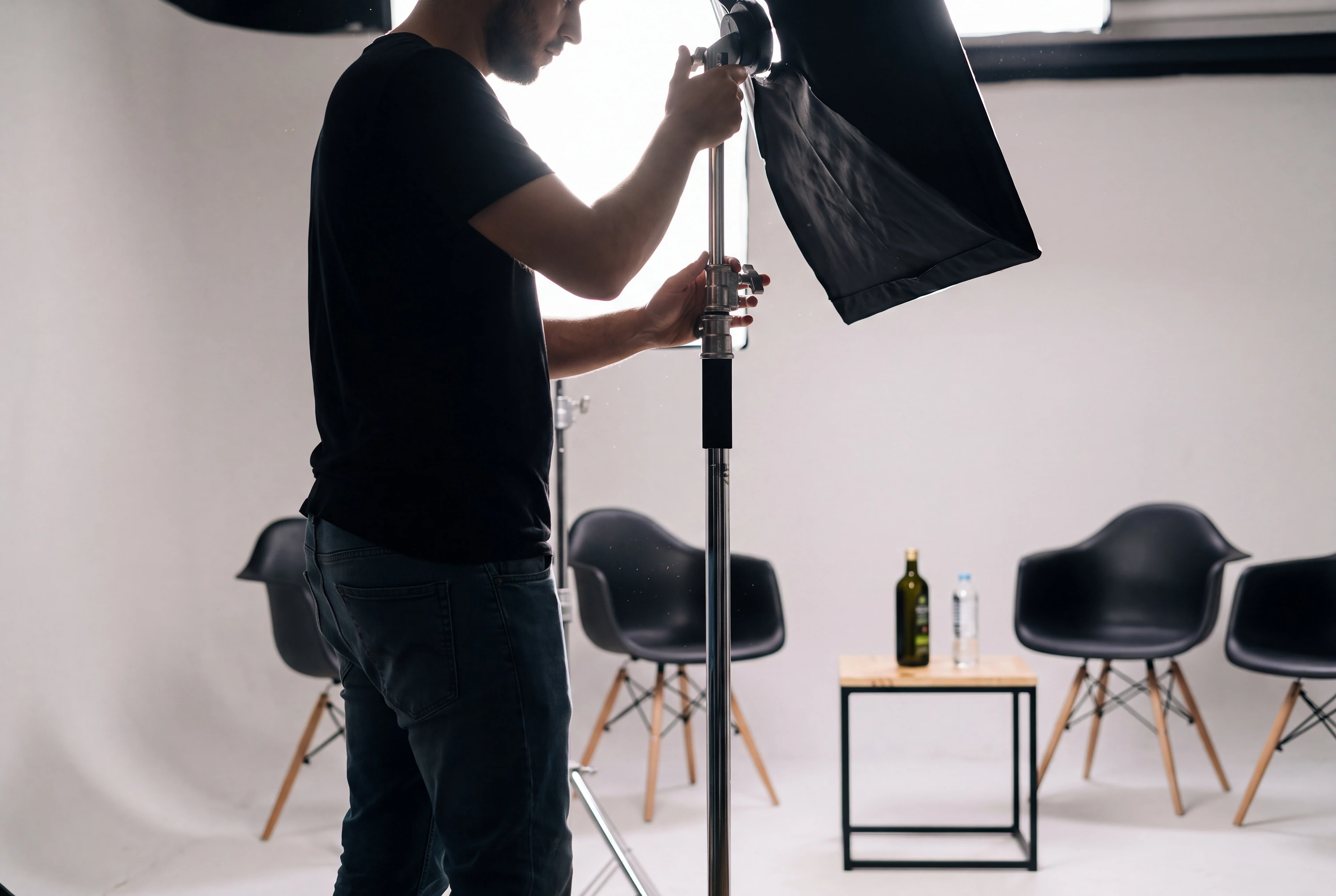 Um técnico ajusta um softbox no estúdio branco, com a mesa de pinho e as cadeiras pretas ao fundo.