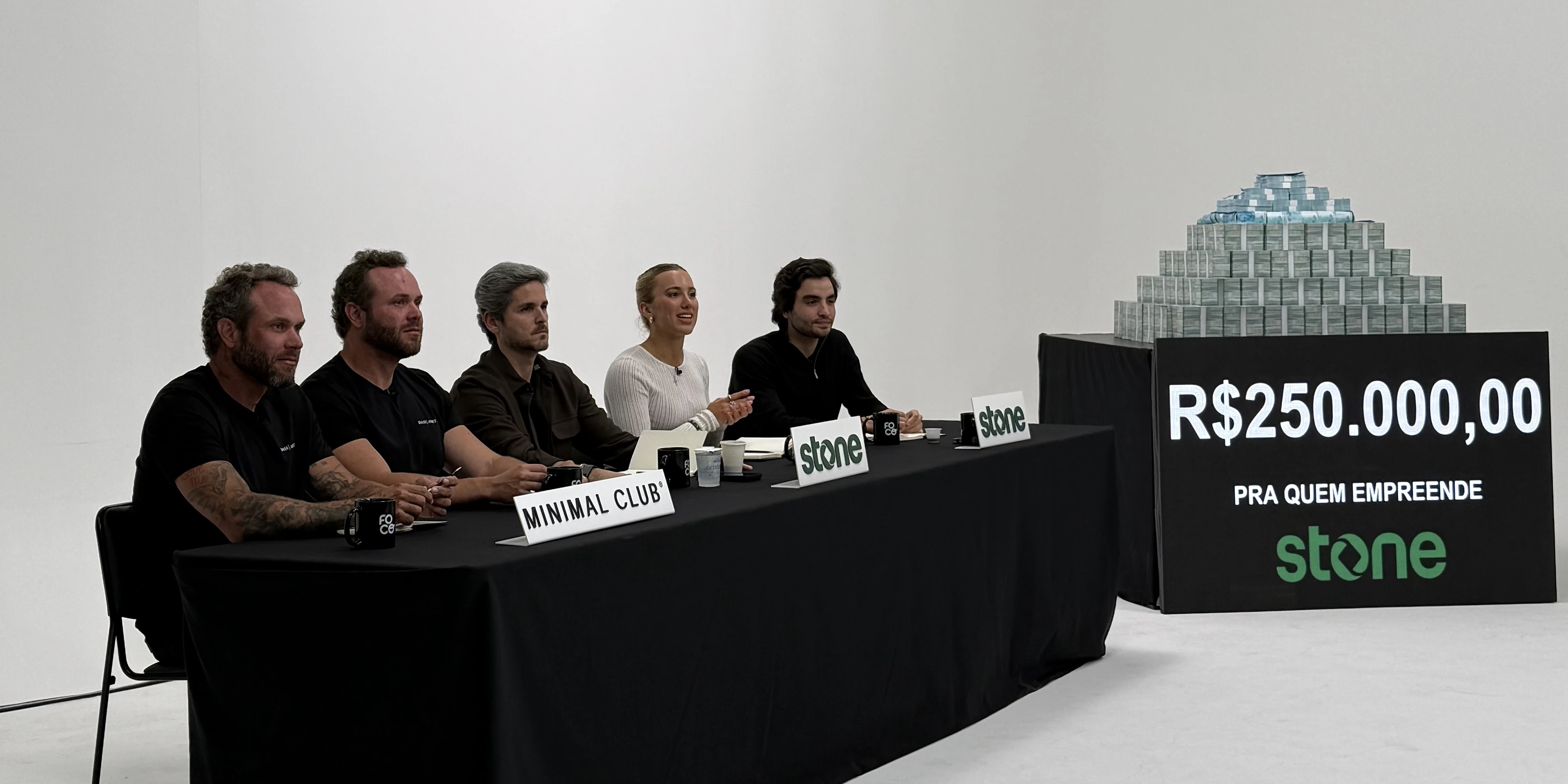 A bancada de Os Caras do Pix atrás da mesa preta, com as placas da Minimal Club e da Stone, e a pilha de dinheiro do prêmio de R$ 250 mil ao lado.